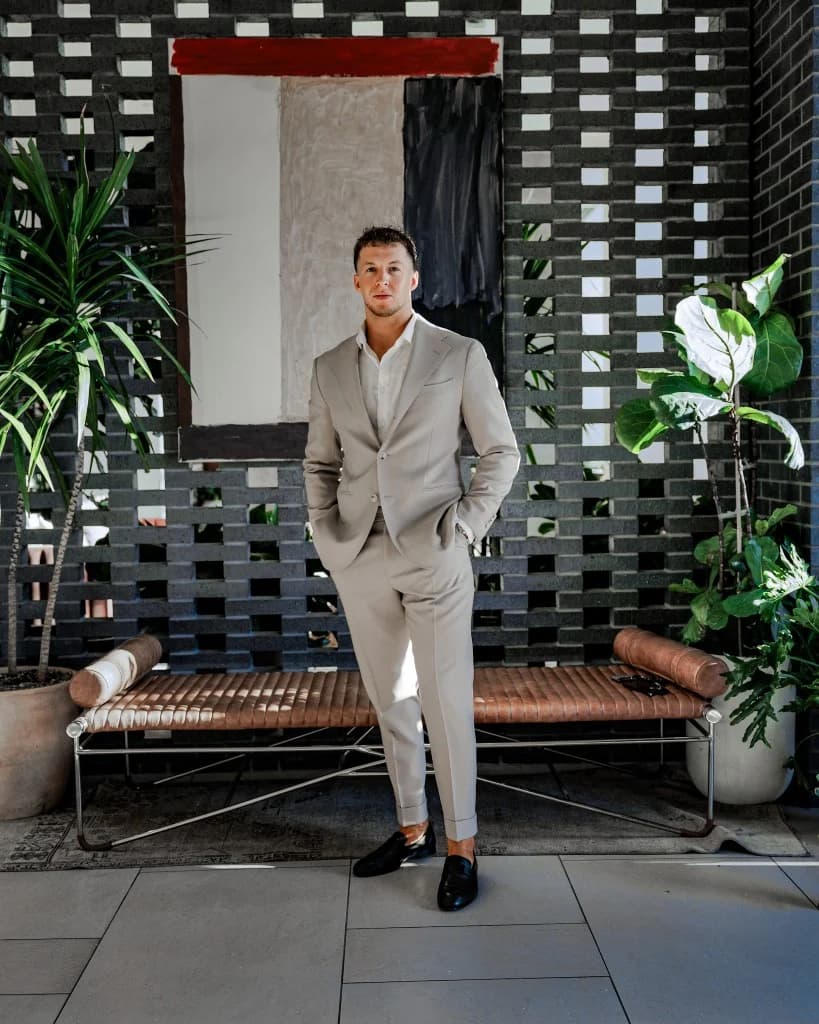 Hayden Mitchell, founder of RevCore HQ. Editorial portrait in a tailored light beige suit over an open-collar white shirt and black loafers, hands in pockets, standing in front of a red-white-black abstract painting on a perforated black brick wall flanked by tropical plants.